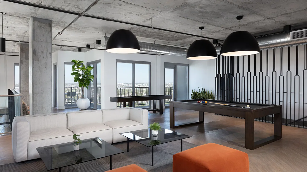 Modern lounge with a black pool table, white sofa, orange ottoman, glass coffee table, and large hanging lights.