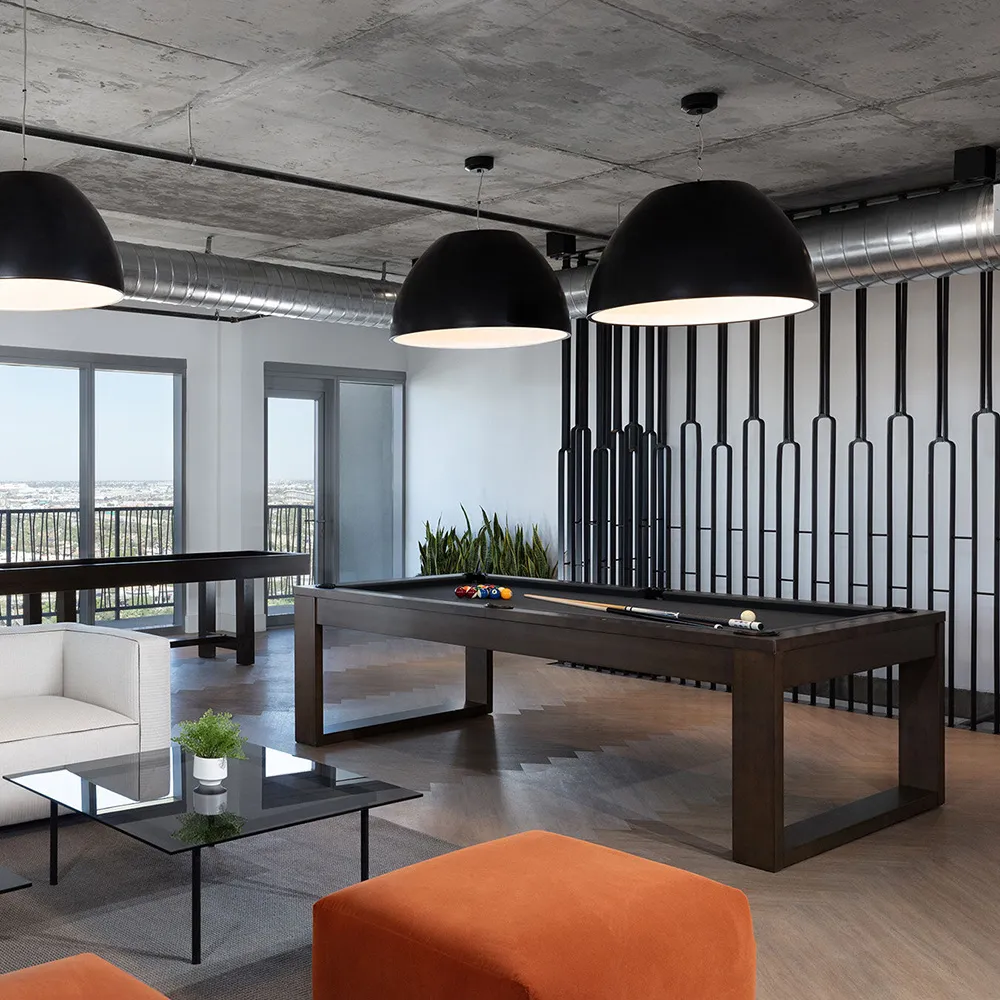 Modern lounge with a black pool table, white sofa, orange ottoman, glass coffee table, and large hanging lights.
