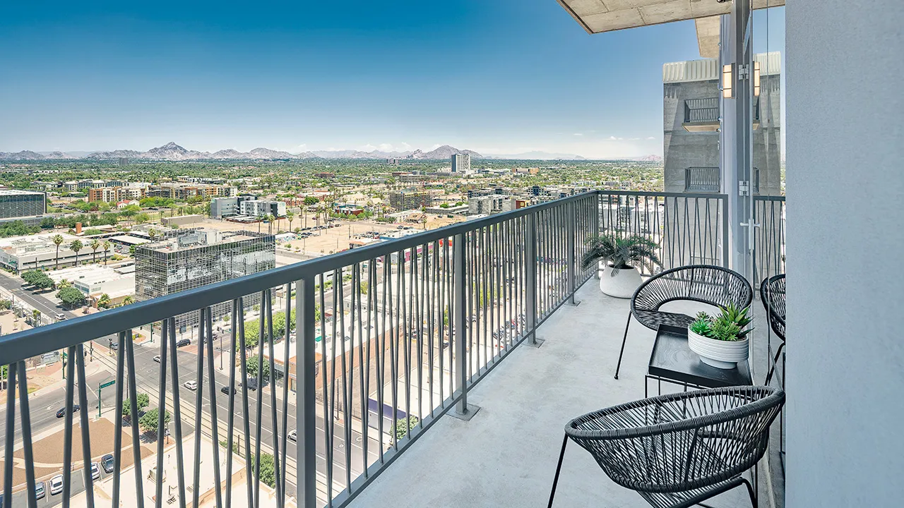 High balcony with two black chairs, table with a plant, overlooking a city and distant mountains.