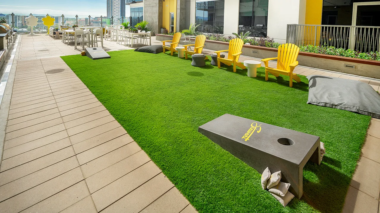 Outdoor courtyard with green grass, yellow Adirondack chairs, tables, and cornhole game boards with bags.