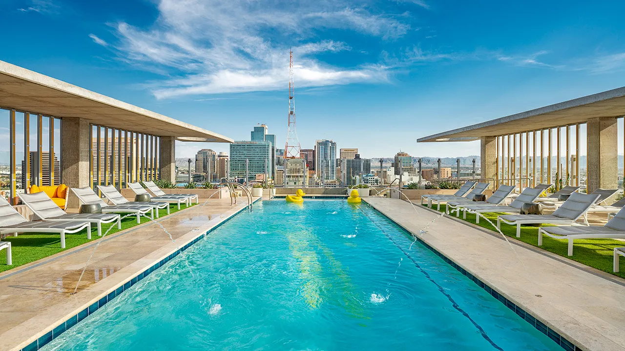 Rooftop pool with water fountains, yellow duck floats, white lounge chairs, and city skyline in background.