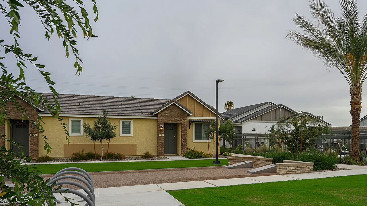 Single-story residential homes with yellow and white exteriors, green lawns, and palm trees on a cloudy day.