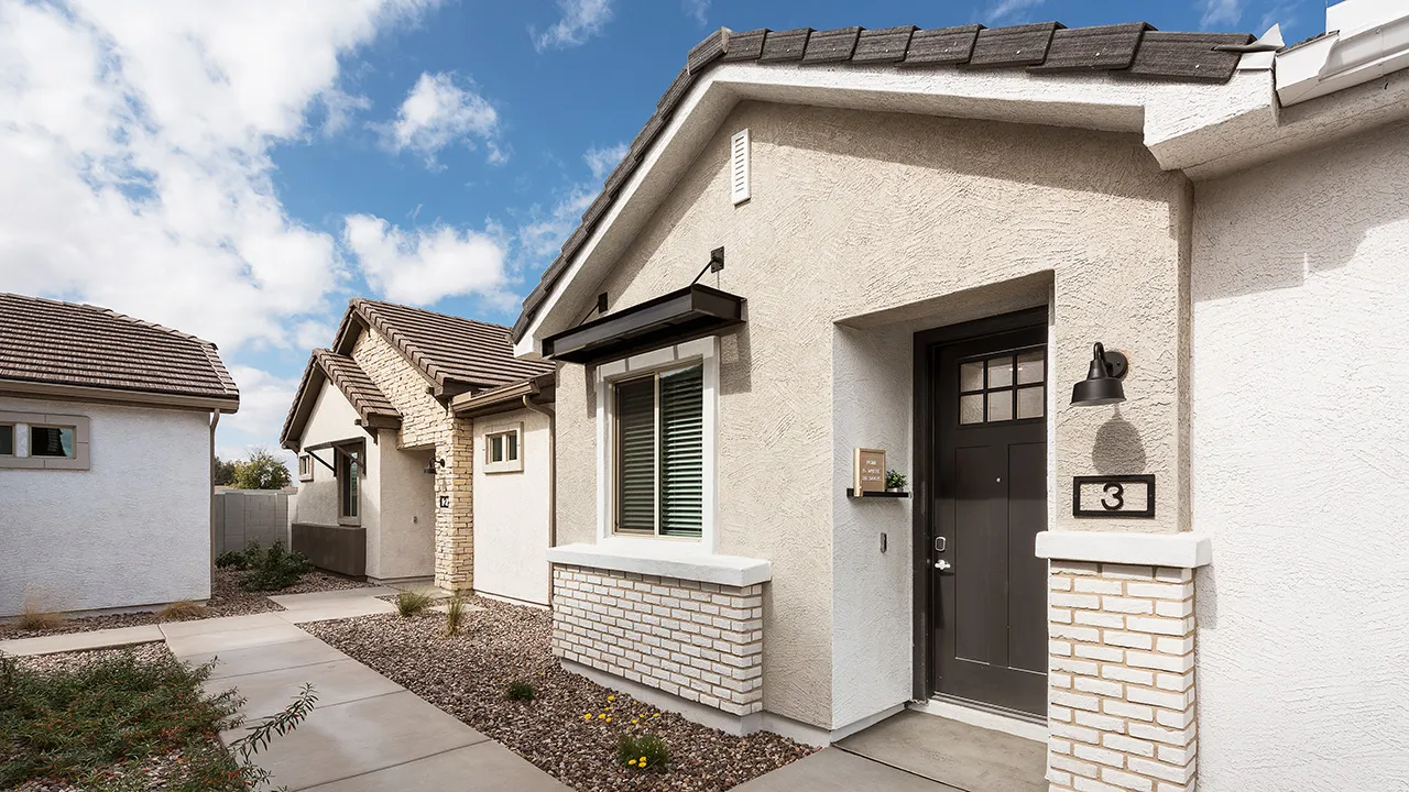 Modern beige stucco houses with dark doors, tiled roofs, and small front yards under a partly cloudy sky.