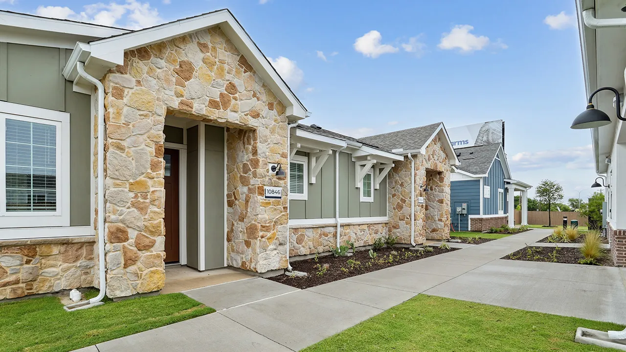 Stone and siding townhomes with covered entryways along a concrete walkway under a blue sky.