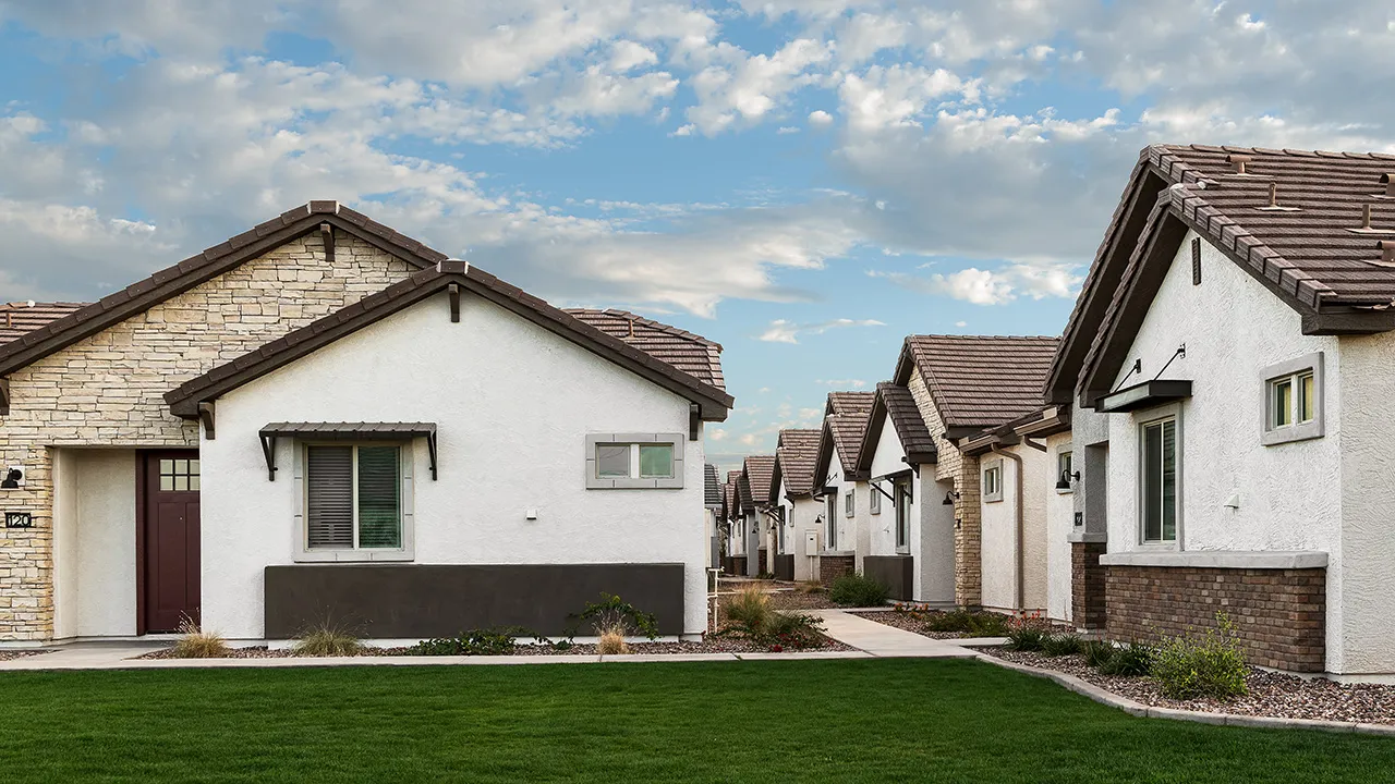 Row of modern single-story houses with white walls, stone accents, and green lawns under a partly cloudy sky.