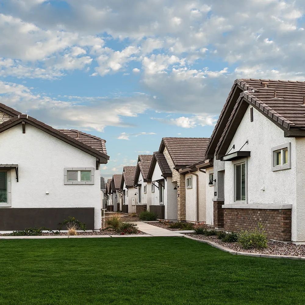 Row of modern single-story houses with white walls, stone accents, and green lawns under a partly cloudy sky.