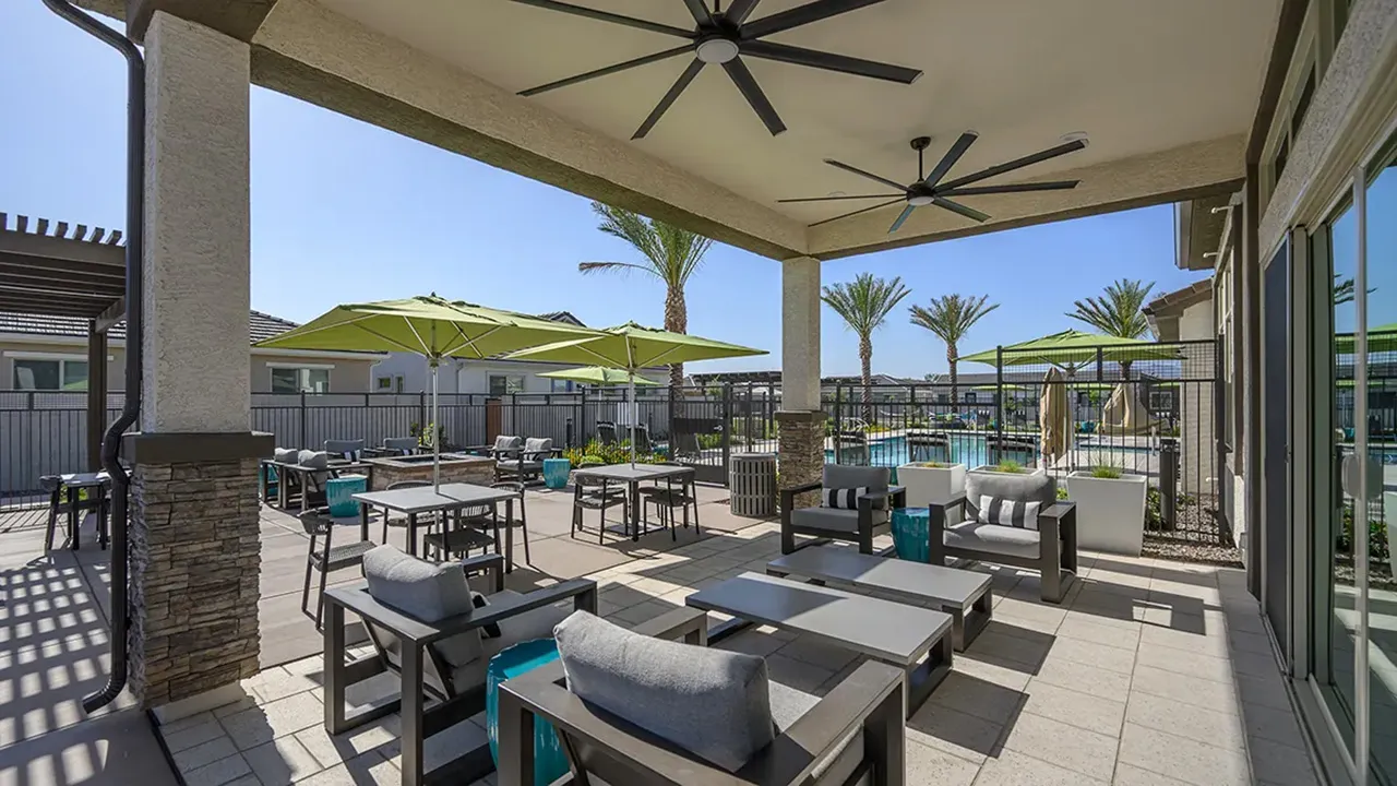 Outdoor patio with cushioned chairs, tables, green umbrellas, ceiling fans, palm trees, and a swimming pool.