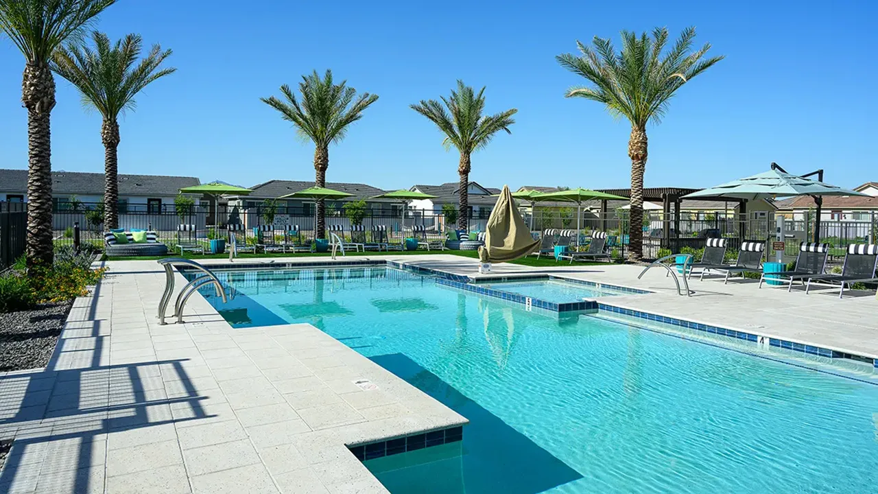 Clear blue swimming pool with palm trees, lounge chairs, and green umbrellas under a sunny sky.