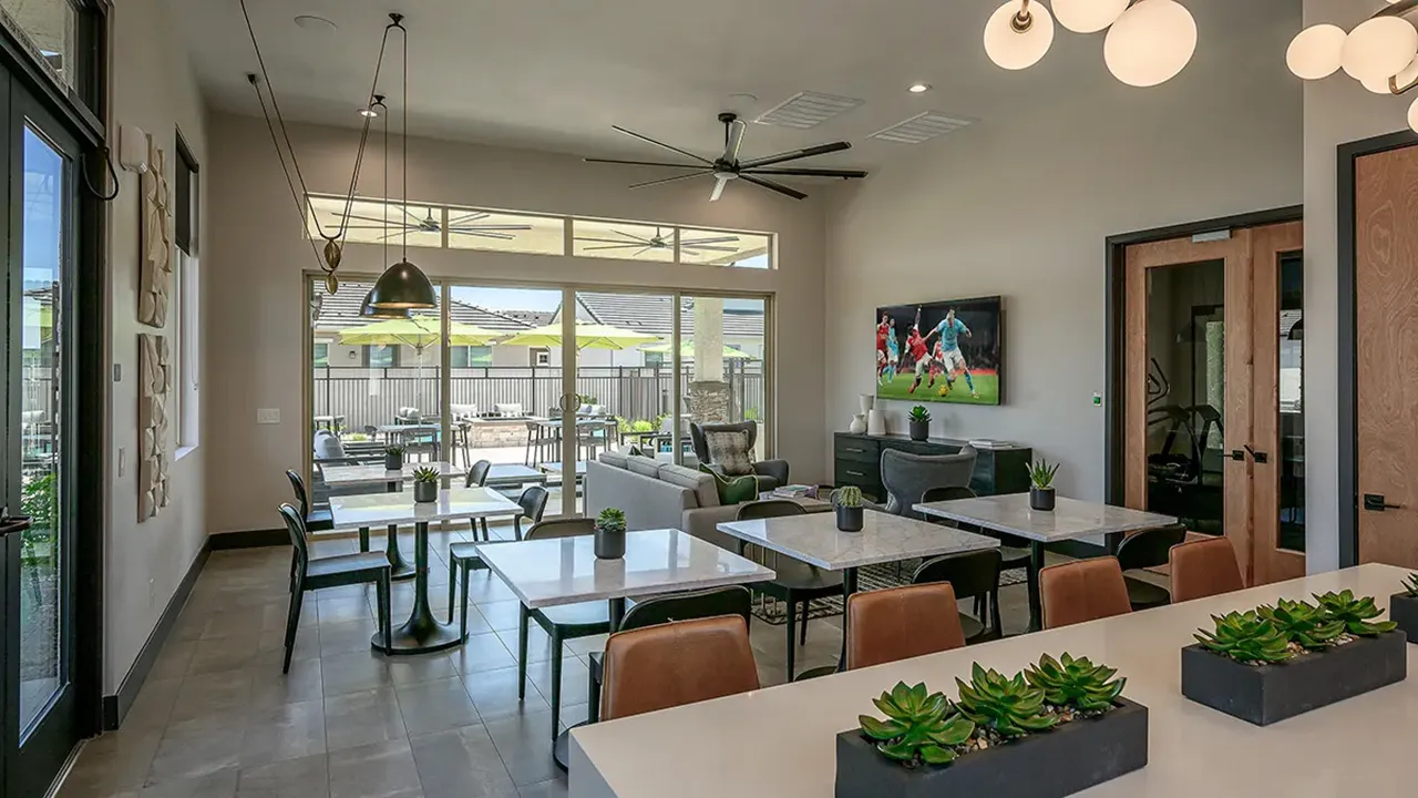 Modern clubhouse interior with seating area, kitchen, tables, chairs, and large ceiling fan.