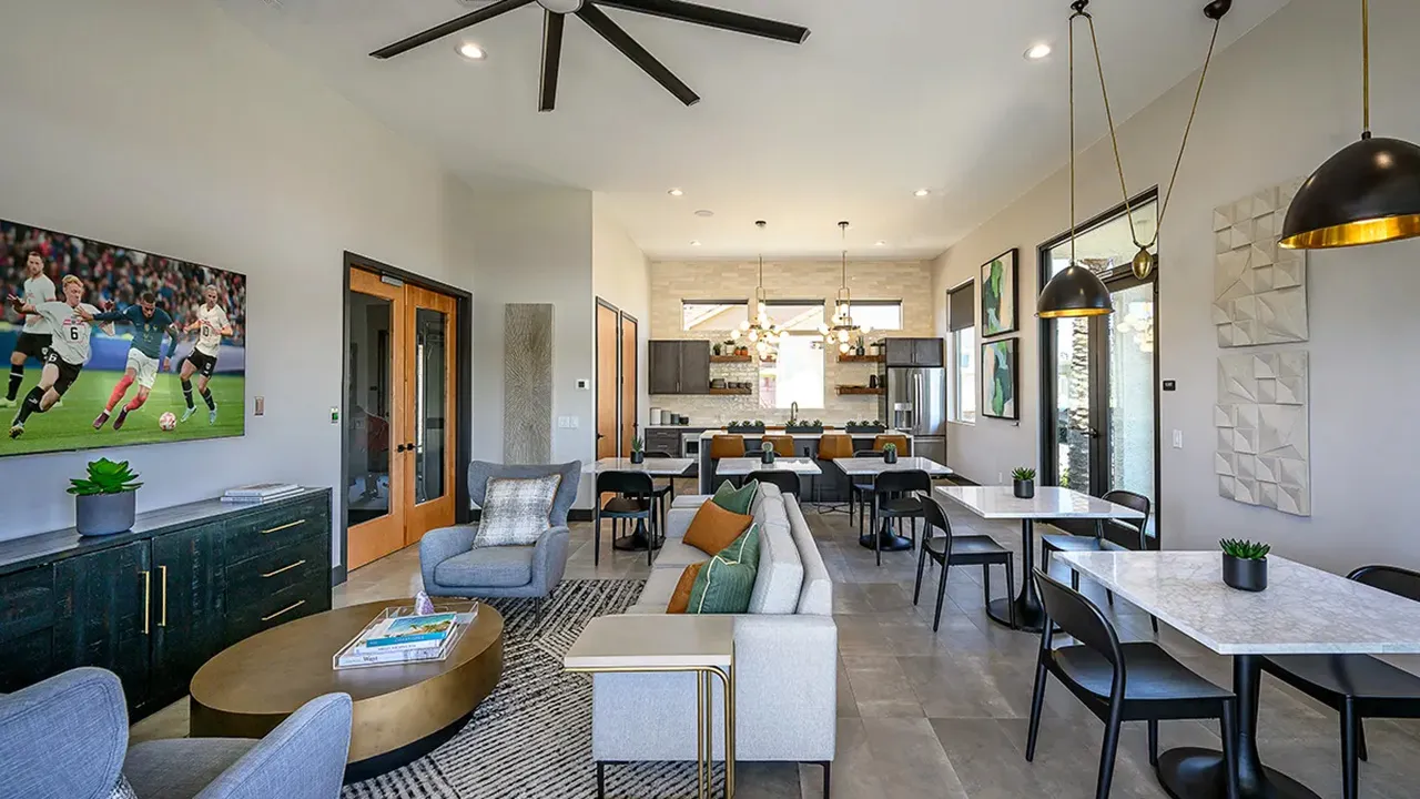 Modern clubhouse interior with seating area, kitchen, tables, chairs, and large ceiling fan.