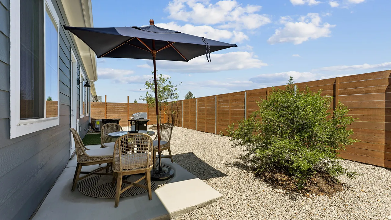 Patio with round table, wicker chairs, a large dark umbrella, and a gravel yard with wooden fence.