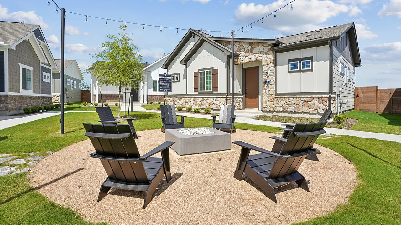 Outdoor fire pit area with six black chairs on gravel surrounded by grass and houses.