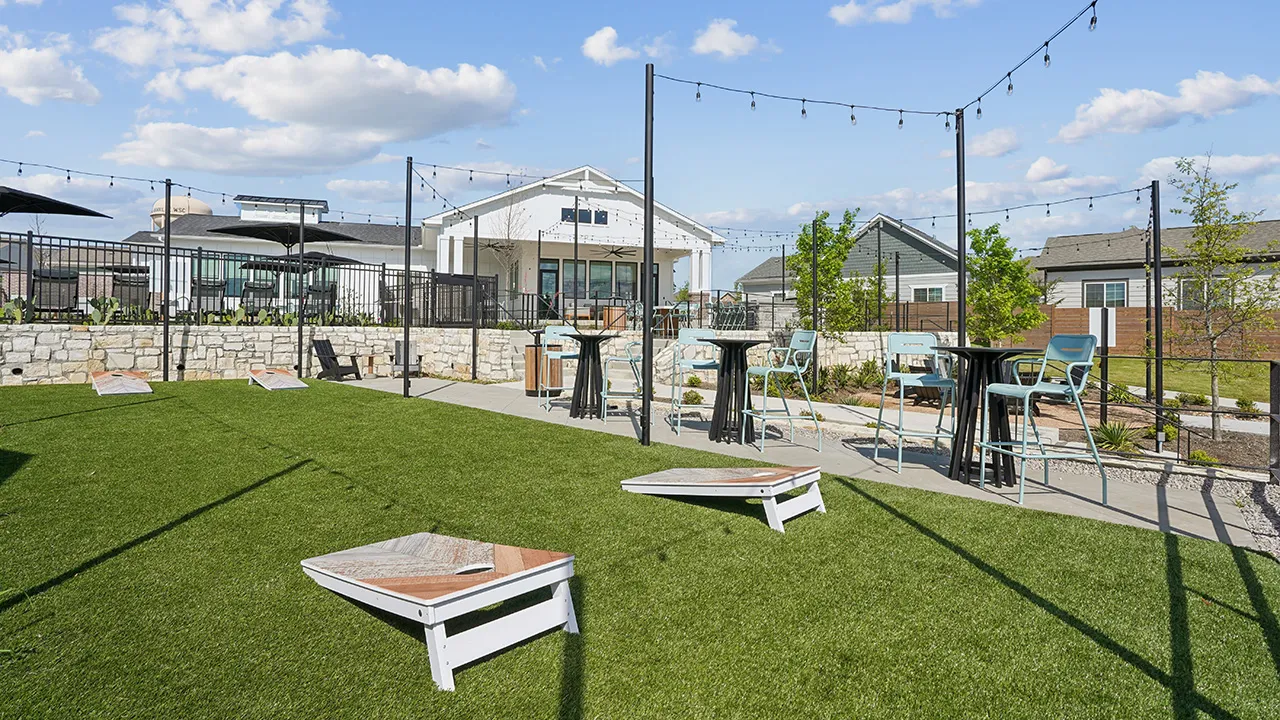 Outdoor lawn area with cornhole boards, string lights, tables, and chairs by residential buildings.