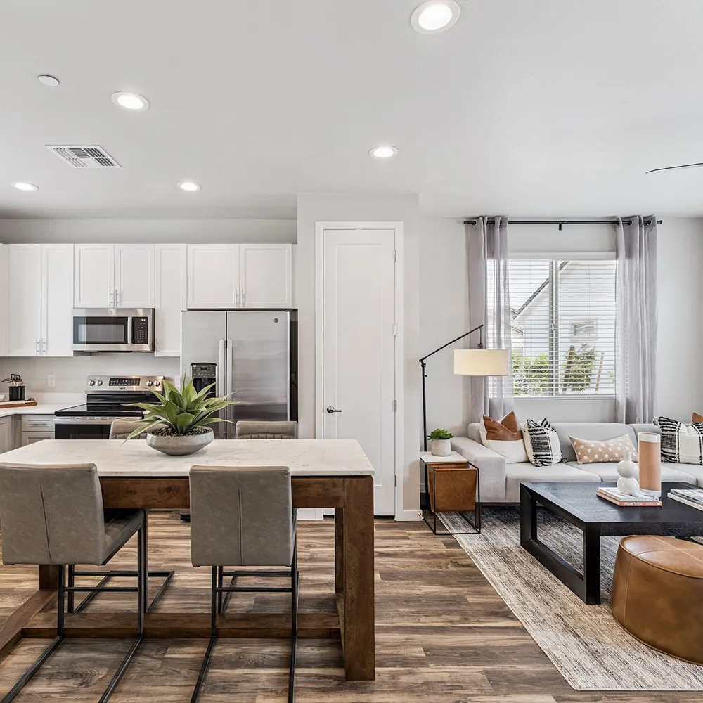 Modern living room with gray sectional sofa, leather stools, dark coffee table, and kitchen in background.