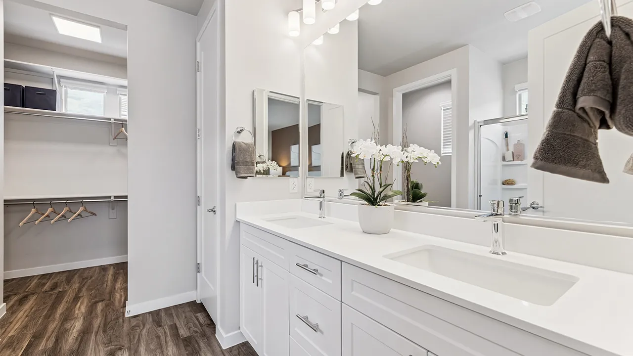 Modern bathroom with white double sink vanity, large mirror, orchid plant, and walk-in closet with wood floor.
