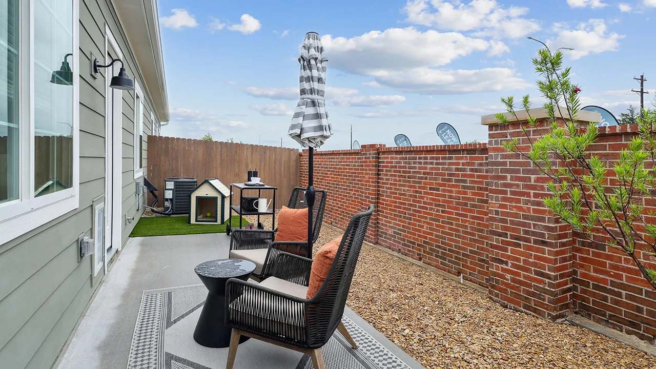 Outdoor patio with two cushioned chairs, small table, closed umbrella, doghouse, and brick wall under blue sky.