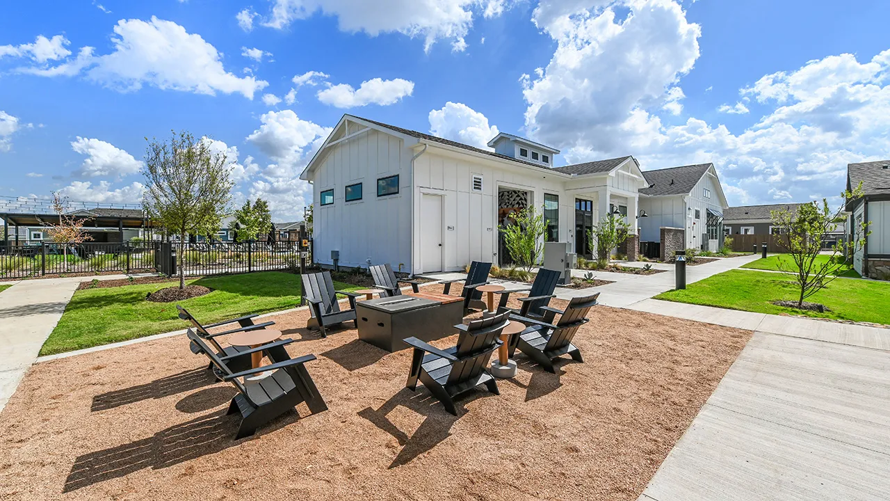 Black chairs arranged around a firepit on gravel outside modern white buildings on a sunny day.