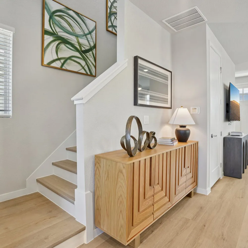 Light wood staircase and cabinet with lamp, sculptures, and art in a modern white home entryway.