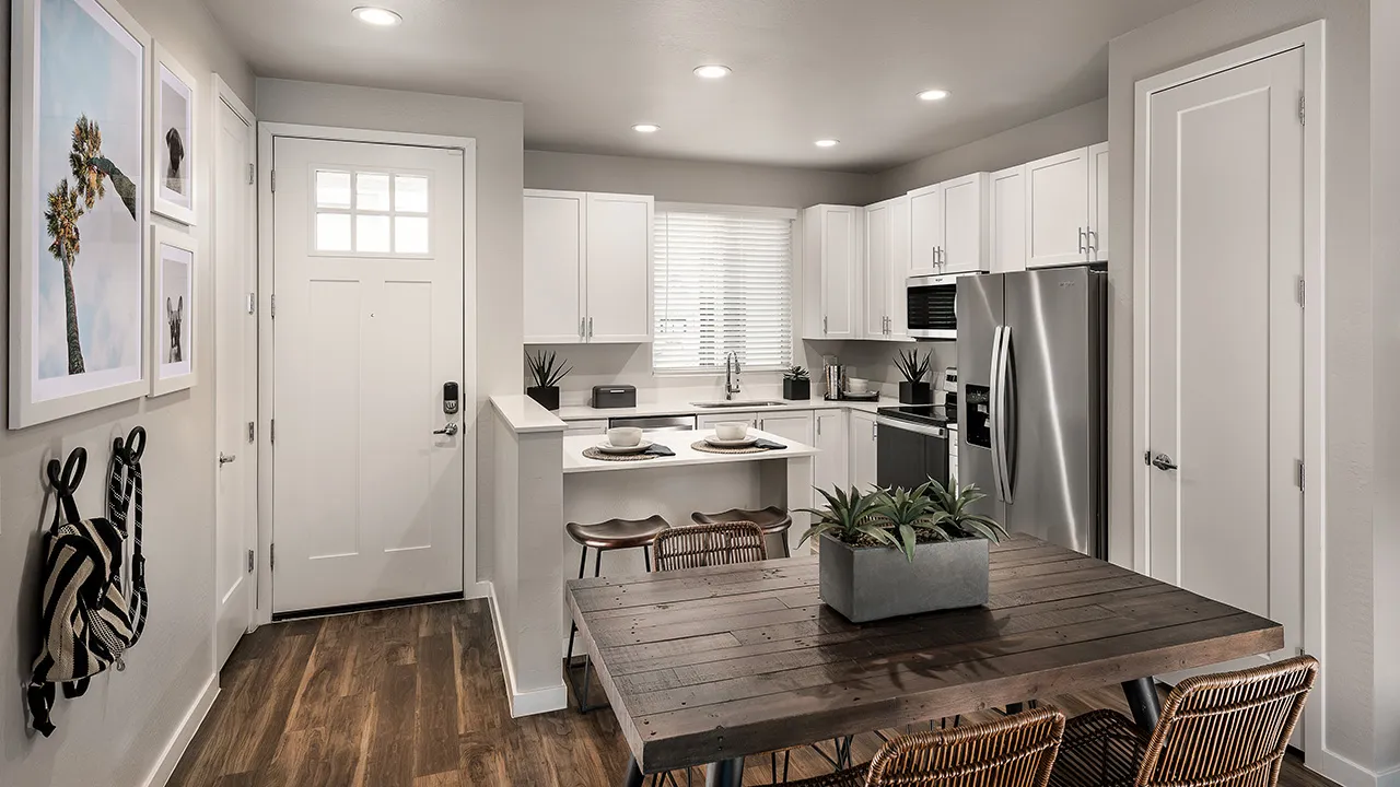 Modern kitchen with white cabinets, stainless steel appliances, wood dining table, and entry door with wall art.