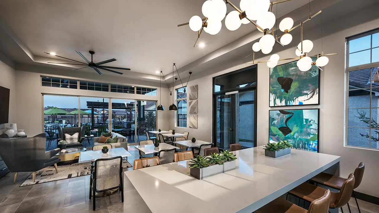 Modern clubhouse interior with seating areas, chandeliers, wall art, and large windows overlooking outdoor patio.