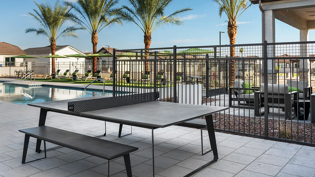 Modern outdoor ping pong table with bench by pool and palm trees under blue sky.