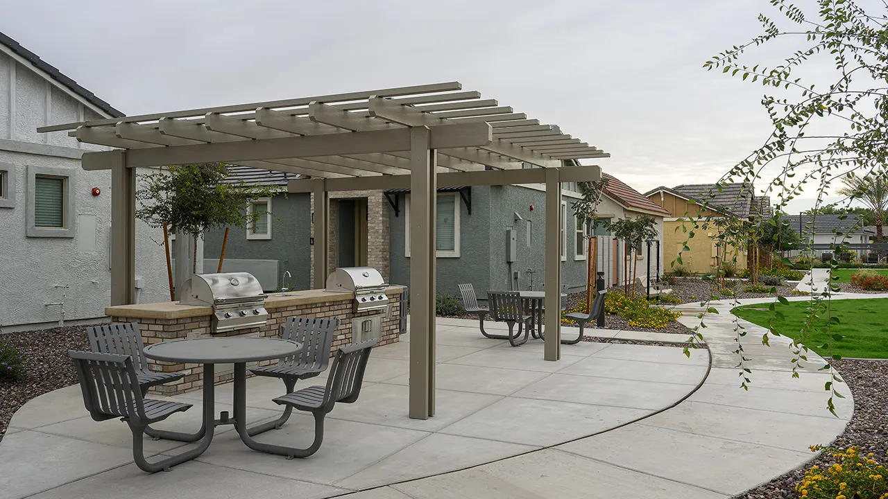 Community outdoor grill area with two built-in grills, shaded picnic tables, and nearby houses.