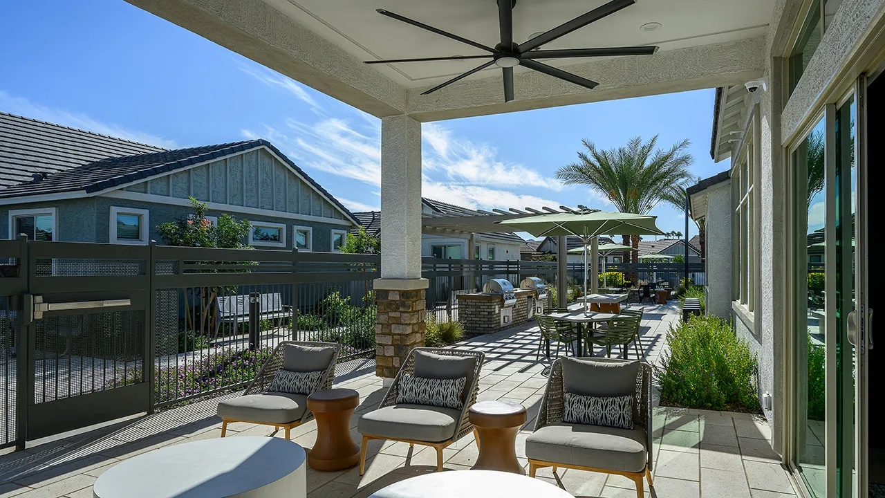 Outdoor patio with modern chairs, tables, large ceiling fan, umbrellas, and built-in grills in a sunny setting.