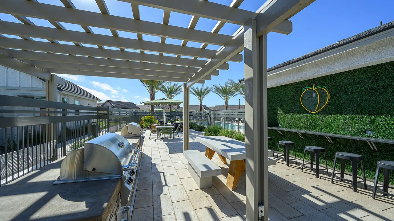 Outdoor patio with grills, picnic table, seating under pergola, palm trees, and a pool in the background.