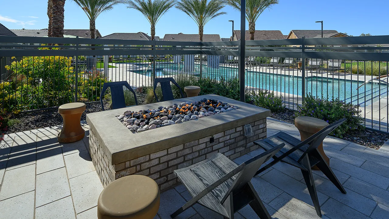 Outdoor fire pit with black chairs and stools next to a fenced swimming pool on a sunny day.