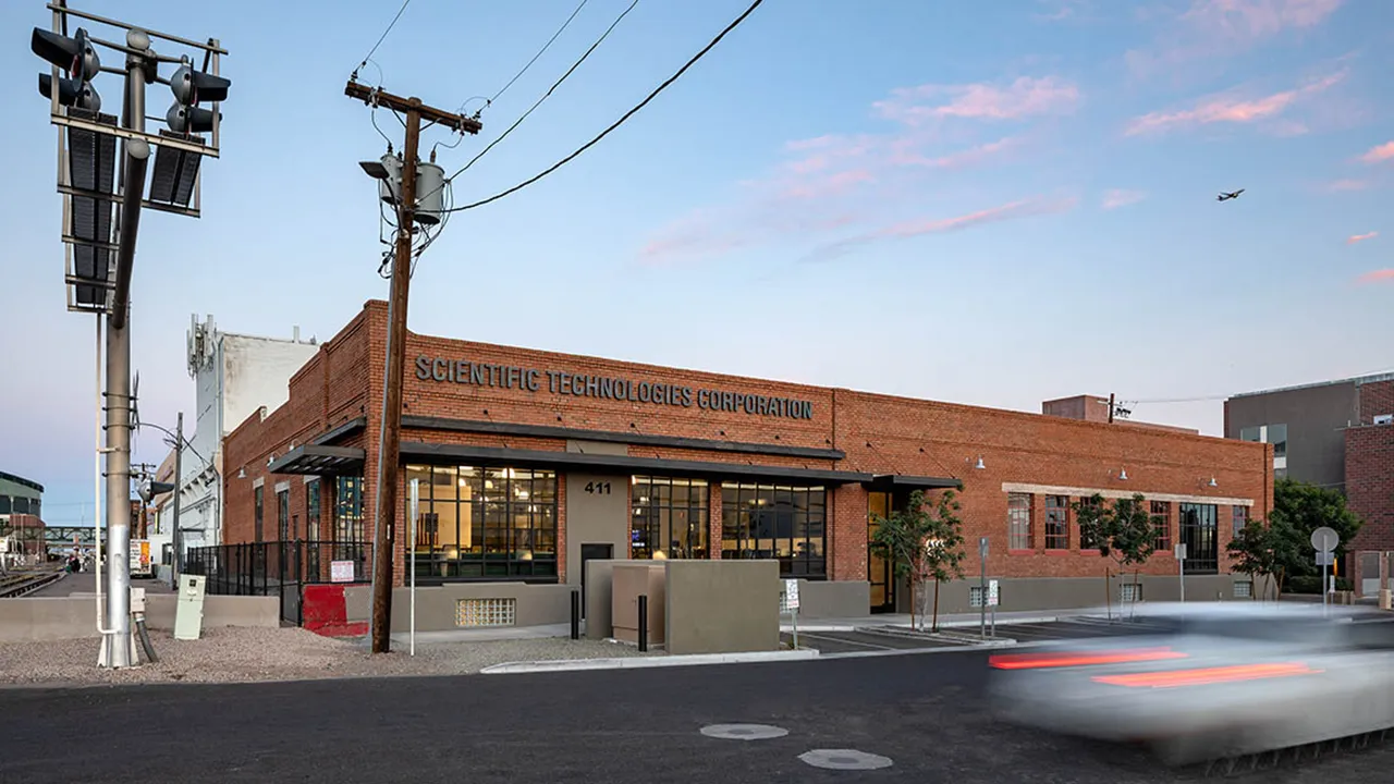 Brick building with large windows labeled 'Scientific Technologies Corporation' at address 411, next to a railway crossing with signal lights and a passing blurred car on the street at dusk.
