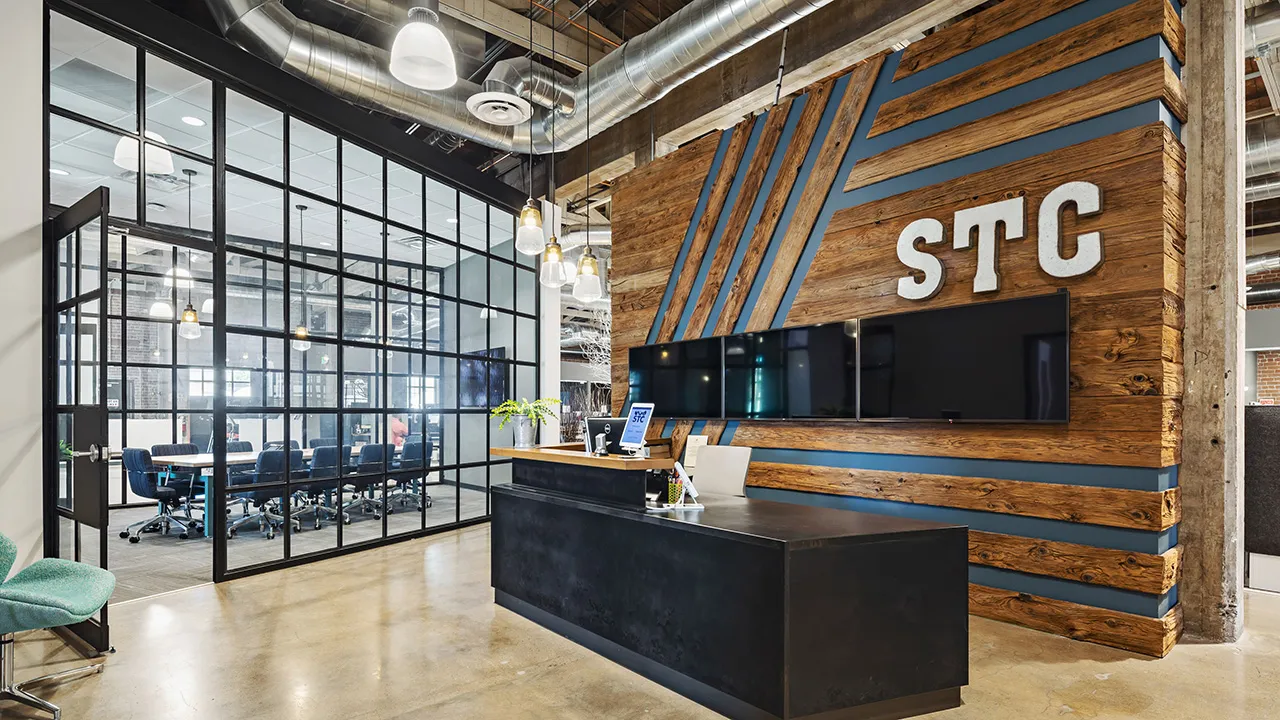 Modern office reception area with a black desk, wood-paneled wall featuring blue diagonal stripes and large white letters 'STC', three flat screen monitors mounted on the wall, and a glass-walled conference room with blue chairs visible through the glass.