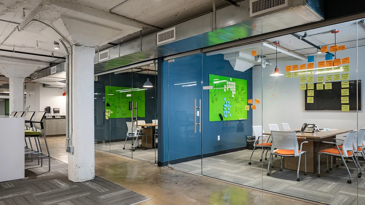 Modern office breakout rooms with a large table surrounded by chairs, a wall-mounted flat screen TV, hanging pendant lights, and glass walls showing an open office area with desks, chairs, and exposed ceiling pipes.