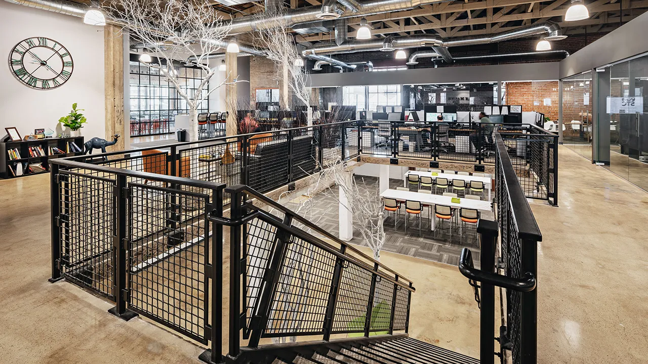 View from the top of stairs of a spacious modern industrial office area with exposed ceiling and brick walls and wooden ceiling beams.