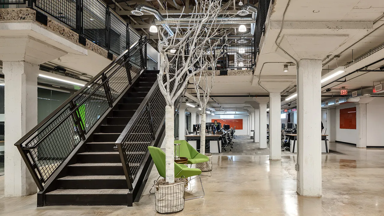 The bottom of a black industrial style staircase of a modern office with white pillars and many workspaces in the distance. 