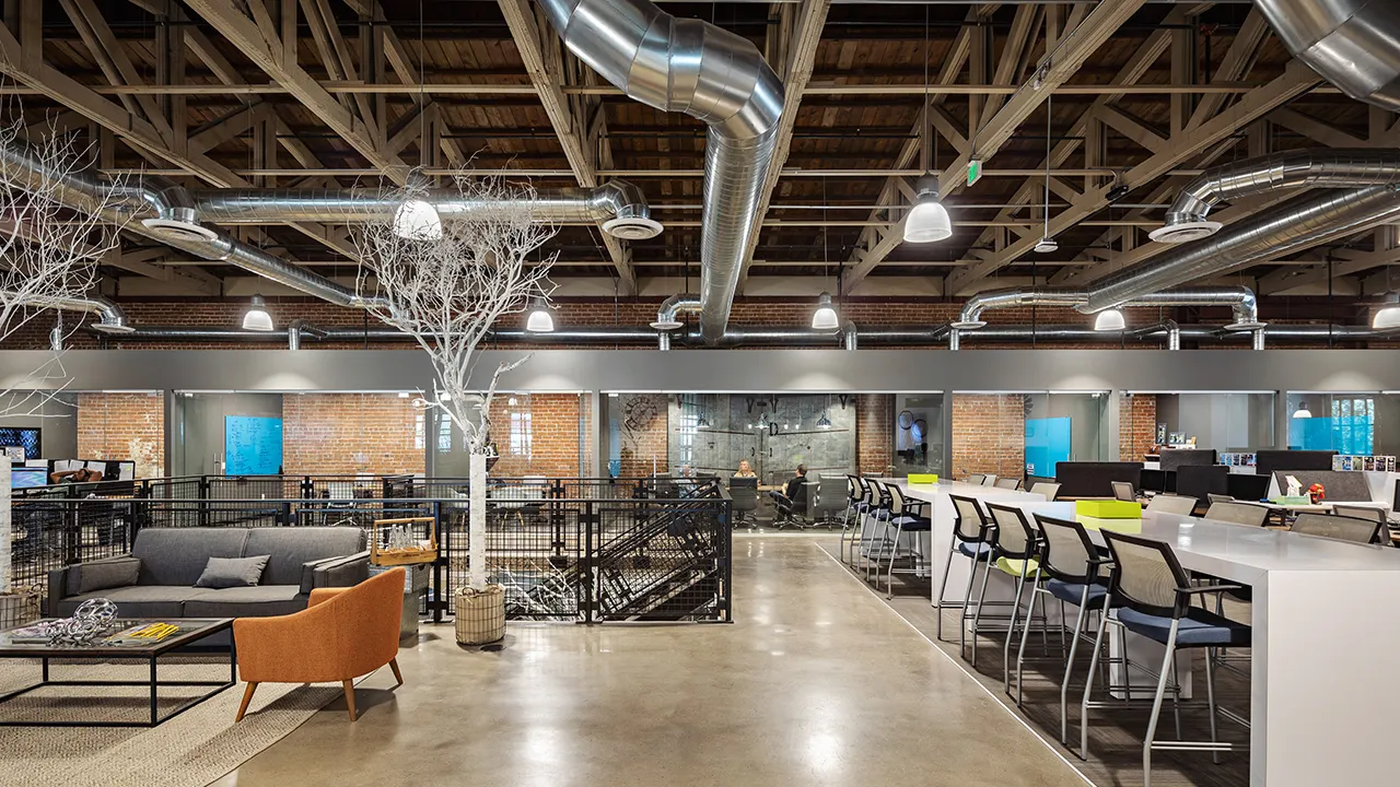 Spacious modern industrial co-working area with exposed ceiling and brick walls and wooden ceiling beams, featuring white waterfall counters surrounded by chairs.