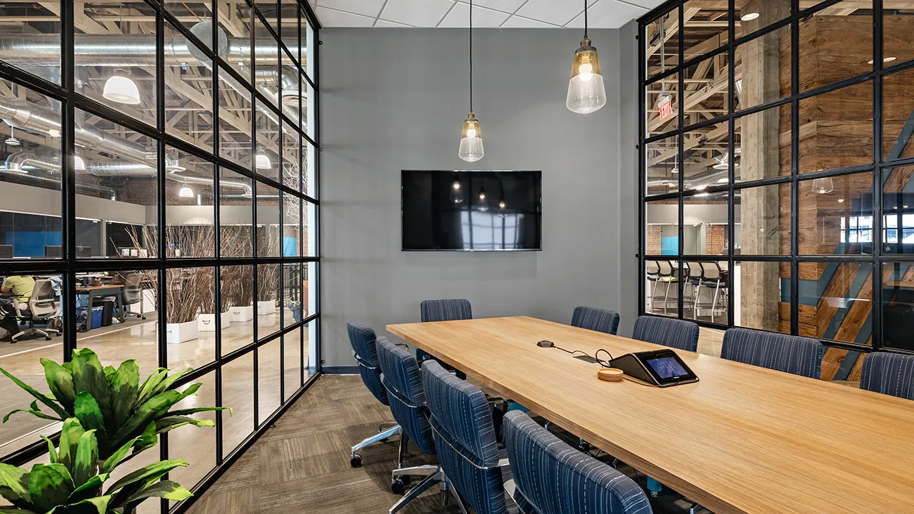 Modern conference room with a long wooden table surrounded by eight blue upholstered chairs, a wall-mounted flat screen TV, two hanging pendant lights, and glass walls showing an open office area with desks, chairs, and exposed ceiling pipes.