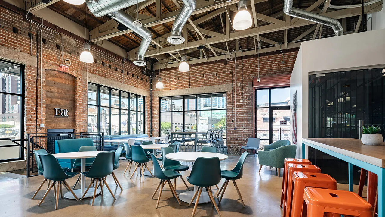 Spacious modern industrial dining area with exposed brick walls and wooden ceiling beams, featuring round white tables surrounded by teal chairs, a high table with stools in the background, bright orange stools at a wooden counter on the right, and large windows letting in natural light.