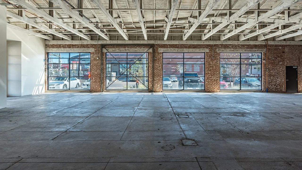 Spacious empty warehouse interior with concrete floor, exposed beams on ceiling, brick walls, and large windows showing parked cars outside.