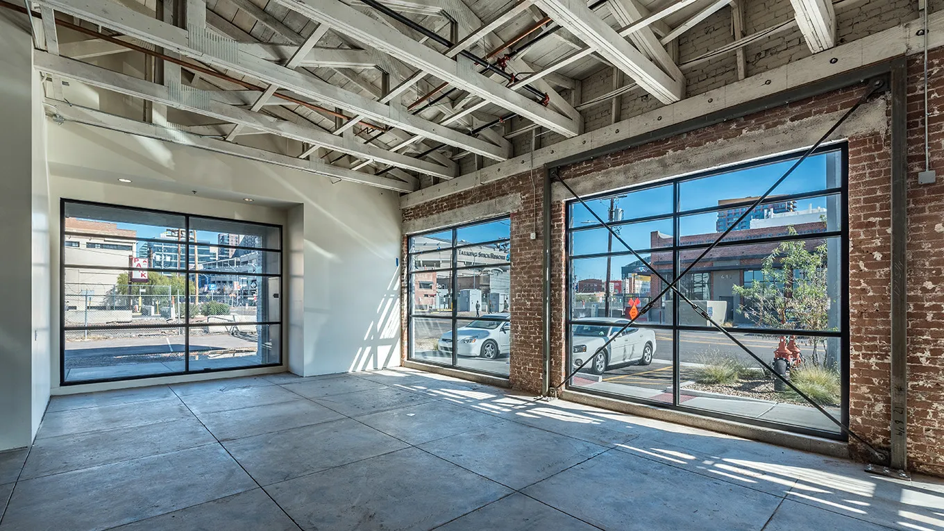 Angled view of spacious empty interior with concrete floor, exposed beams on ceiling, brick walls, and large windows showing parked cars outside.