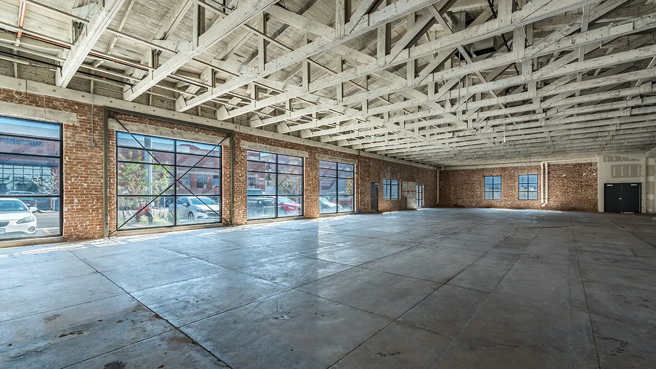 Angled view of spacious empty warehouse interior with concrete floor, exposed beams on ceiling, brick walls, and large windows showing parked cars outside.