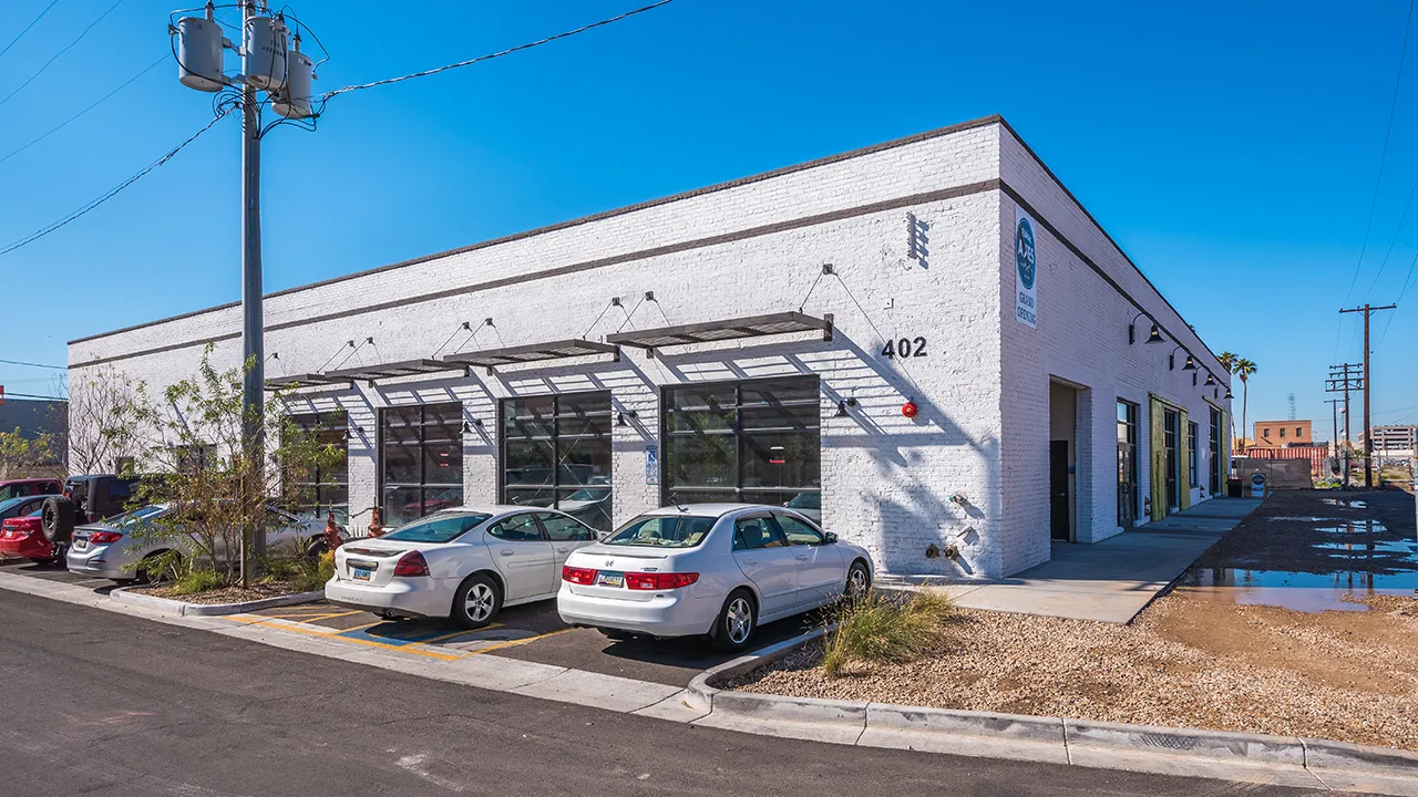 White one-story commercial warehouse building, several parked cars in front, and a utility pole with transformers on the sidewalk.