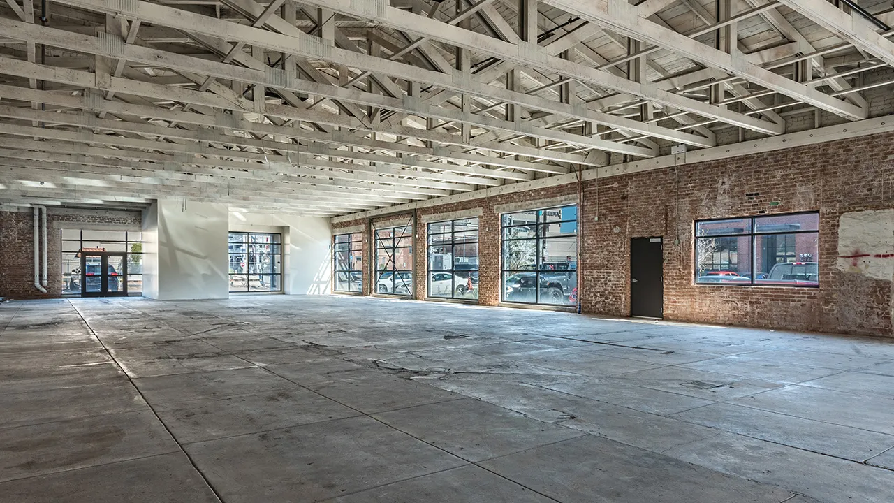Angled view of spacious empty warehouse interior with concrete floor, exposed beams on ceiling, brick walls, and large windows showing parked cars outside.