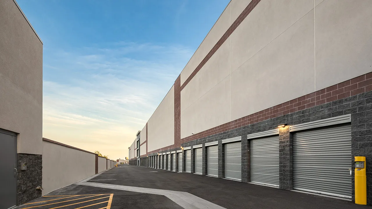 Exterior view of a self-storage facility with multiple closed roll-up garage doors along a paved driveway, under a clear blue sky.
