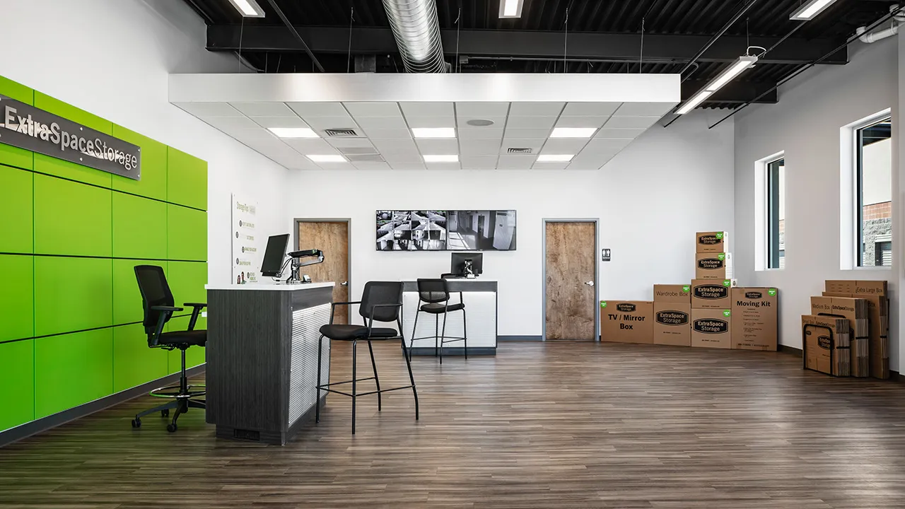 Modern storage office interior with a green accent wall labeled Extra Space Storage, a service desk with chairs, security monitor screens on the back wall, two wooden doors, and a stack of labeled moving and storage boxes in the corner.
