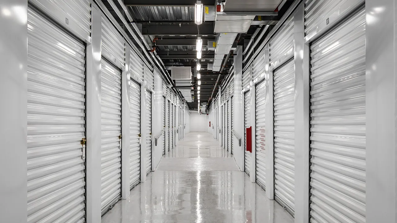 Long hallway of indoor storage units with closed white roll-up doors on both sides, shiny polished floor, and fluorescent ceiling lights.