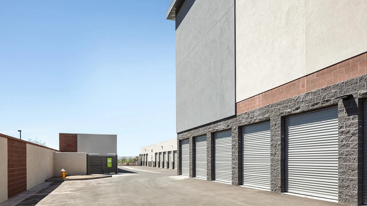 Exterior view of a self-storage facility with multiple closed roll-up garage doors along a paved driveway, under a clear blue sky.