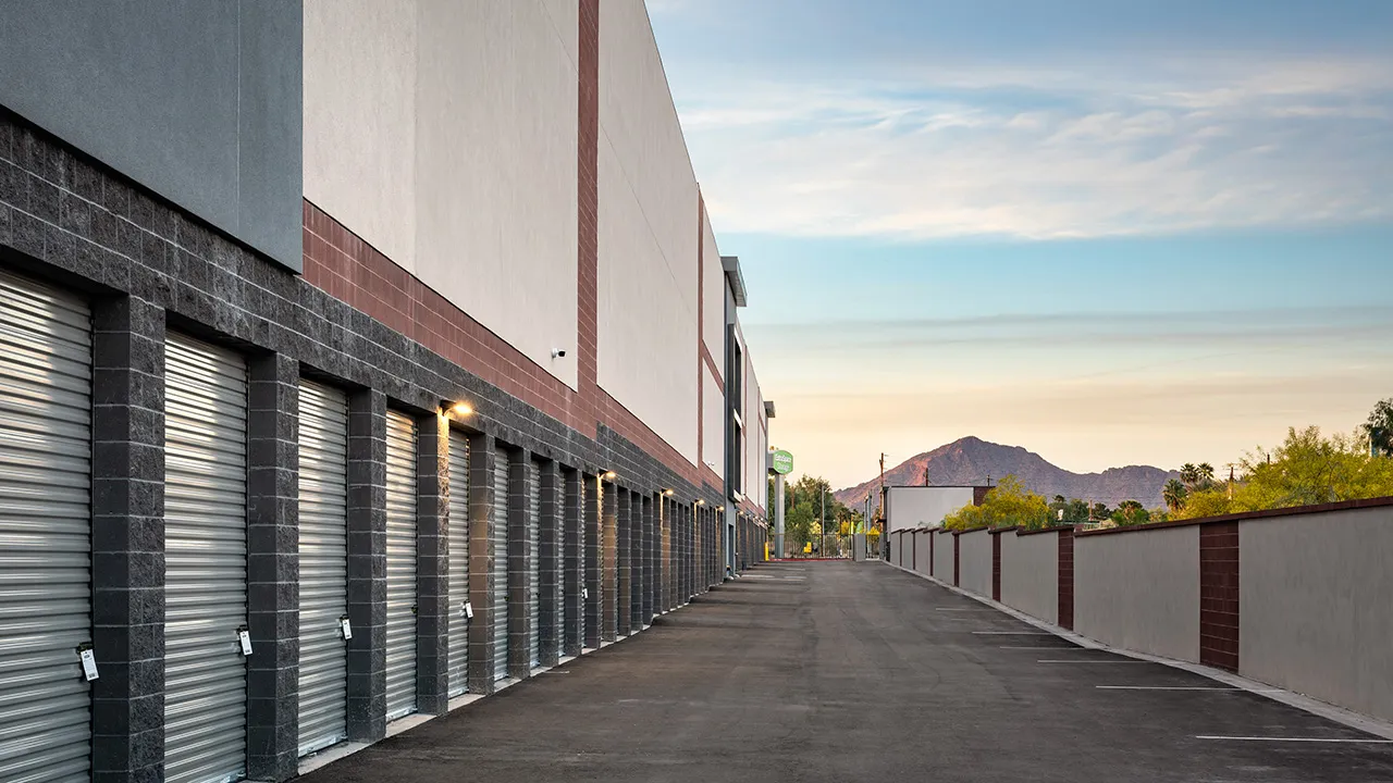 Exterior view of a self-storage facility with multiple closed roll-up garage doors along a paved driveway with a desert mountain in the background at sunet.