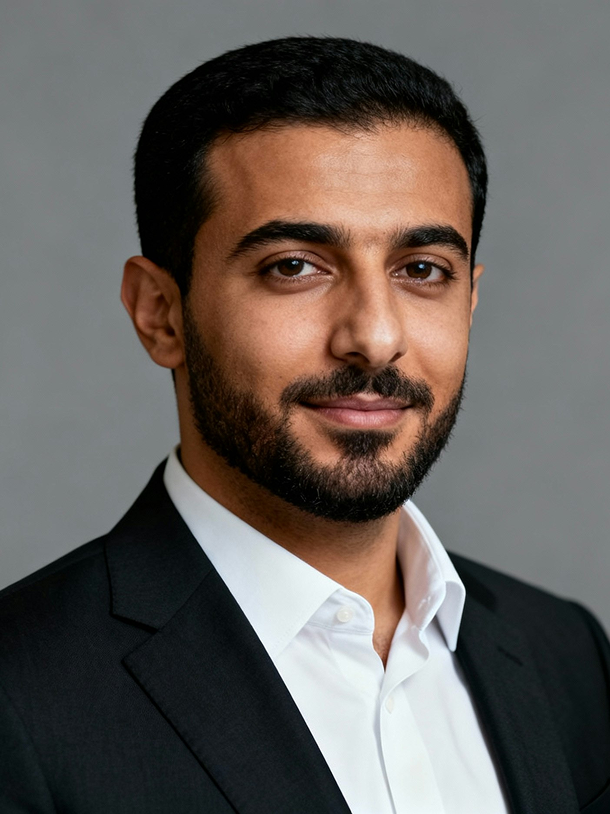 Professional headshot of a man with dark hair and beard wearing a black suit jacket and white dress shirt against a gray background.