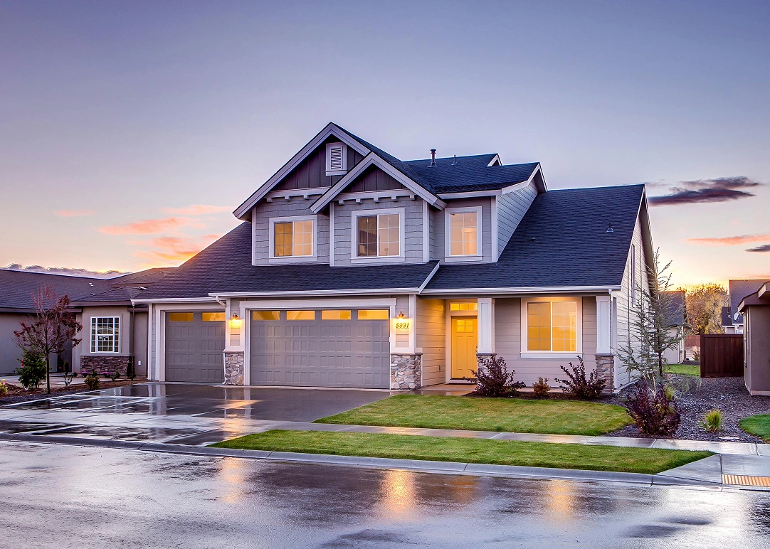 Modern two-story suburban house with multiple garage doors, illuminated windows, and wet driveway at dusk.