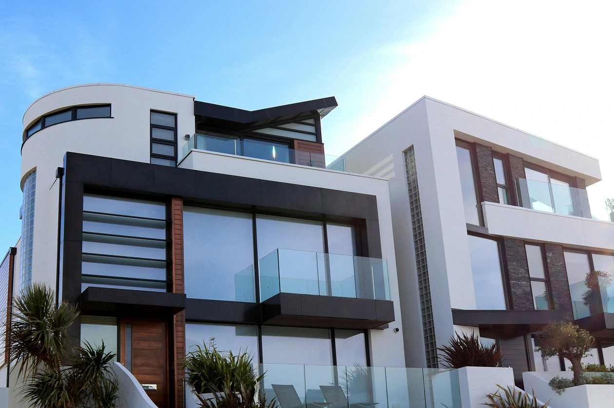 Modern multi-story house with large glass windows, black framing, balconies, and surrounding greenery under a clear blue sky.